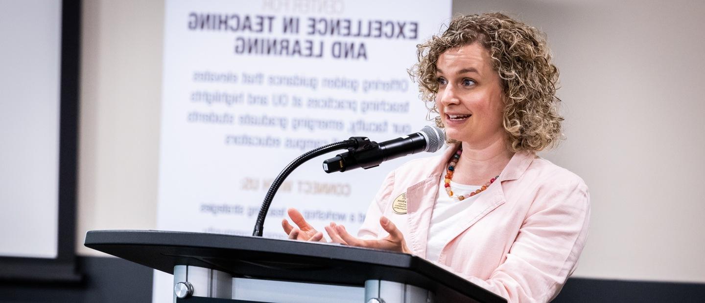 A woman stands at a podium in front of a microphone. In the background a slide is on a screen.